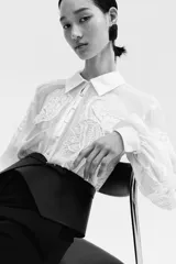 Blusa oversized de tejido transparente color crudo, con detalles bordados en relieve, cuello camisero, cierre frontal de botones y mangas largas abullonadas con puños ajustados.