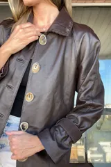 Tapado de cuero marrón con diseño artesanal, cuello camisero con detalles de pasamanería en gamuza, cierre frontal con botones de guampa y bolsillos delanteros con bordes decorativos. Presenta un corte cómodo y versátil.