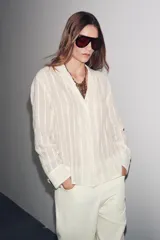 Camisa de algodón color crema o blanco roto, con textura de rayas verticales sutiles. Presenta cuello solapa, escote pico y manga larga.