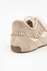 Zapatilla de cuero legítimo en tonos beige y crema, con diseño de flecos en el lateral y costuras decorativas visibles en el contorno. Presenta cordones al tono y suela plana.