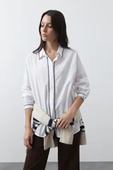 Camisa blanca de manga larga con detalles bordados en contraste a lo largo de la botonadura, el cuello y los puños. Presenta una silueta relajada y un diseño clásico con un toque artesanal.