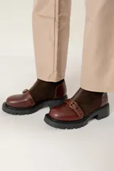 Bota de caña corta combinada en cuero liso y gamuza color marrón. Presenta cierre frontal con cremallera, tira con hebilla metálica decorativa en el empeine y suela track de goma negra.