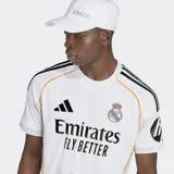 Camiseta de fútbol Adidas del Real Madrid, modelo Primera Equipación temporada 25/26, para hombre. Diseño blanco con detalles en negro (logo Adidas, tres franjas en hombros, texto del sponsor) y ribetes laterales en color naranja/dorado. Incluye escudo del club bordado y logo de sponsor en manga.