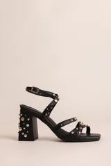 Sandalias de taco ancho de 7 cm, color negro, con tiras cruzadas y cierre de hebilla en el tobillo. El diseño presenta tachas metálicas decorativas en color dorado y plateado distribuidas en las tiras y en todo el taco.