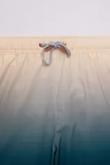 Short de baño con degradé de color beige a azul, con cintura elástica y cordón ajustable. Tiene protección UV50, logo de la marca, dos bolsillos laterales y uno trasero con cierre.