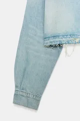 Cazadora corta de denim en tono celeste claro, con cuello solapa, manga larga y cierre frontal con botones. Presenta un bolsillo de plastrón en el delantero y detalles de rotos en el cuello y el ruedo deshilachado.