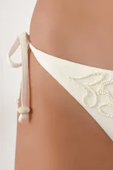 Calzón de bikini de tiro bajo en color crema, con lazos finos ajustables en los costados y detalles de bordado con cuentas en el frente.