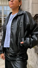 Campera de cuero sintético color negro, con cuello camisero, cierre frontal y costuras visibles en contraste. Presenta un diseño holgado con puños elásticos.