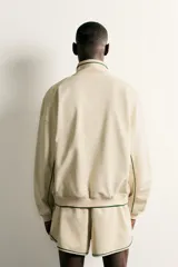 Chaqueta estilo deportivo de corte relajado, color beige con detalles de vivos en color verde oscuro. Presenta cuello alto, cierre frontal con cremallera de doble dirección, bolsillos laterales y puños elásticos.