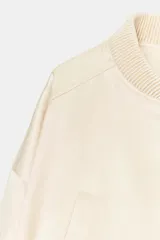 Chaqueta bomber corta de tejido satinado color beige, con cuello redondo, cierre frontal con cremallera, mangas largas de hombro caído y bolsillos de vivo en el delantero. Presenta acabados en rib elástico en cuello, puños y ruedo.