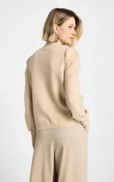 Polera de punto color beige con sutiles destellos brillantes, cuello alto y mangas largas con hombros abullonados y puños ajustados.