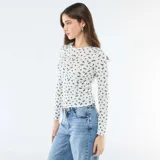 Camiseta de manga larga confeccionada en tejido de punto calado, con estampado de pequeñas flores azules sobre fondo blanco. Presenta un diseño ajustado al cuerpo, cuello redondo y una pequeña abertura frontal con botones.