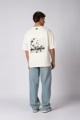 Remera de corte oversize color crudo, con cuello redondo y mangas cortas. Presenta un pequeño estampado gráfico en el pecho con diseño de aves en vuelo.