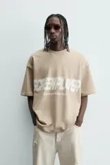 Sudadera de corte relajado, color beige claro, con cuello redondo y manga corta. Presenta un estampado frontal con texto en blanco difuminado que dice "SCREEN PLAYER" y debajo "Hypercore Technology".