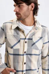 Sobrecamisa de tejido tipo polar con estampado de cuadros en tonos beige, gris y azul. Presenta cuello clásico, cierre frontal con botones oscuros y bolsillos frontales en el pecho.