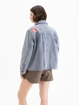 Chaqueta de denim de algodón color azul con efecto lavado, corte recto y cierre frontal con botones. Presenta bolsillos aplicados en el frente y bordados de una abeja y flores.