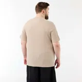 Camiseta de manga corta en color beige, confeccionada en tejido de punto acanalado con sutiles rayas horizontales y cuello redondo. Cuenta con una pequeña etiqueta decorativa en el borde inferior.