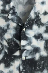 Camisa de manga corta con cuello abierto, tapeta frontal con botones y bajo recto. Presenta un estampado floral con efecto desgastado en tonos blanco y negro.