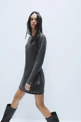 Vestido corto de punto color gris oscuro, con cuello barco y mangas largas acampanadas. Presenta detalles de tachas metálicas aplicadas en el ruedo y en los puños, con un diseño de corte evasé.