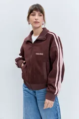 Cazadora de estilo deportivo en color marrón, confeccionada en tejido tipo gamuza. Presenta cierre frontal completo, cuello alto, puños y ruedo acanalados, y franjas laterales en tono rosado. Incluye el logo de la marca bordado en el pecho.