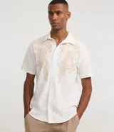 Camisa de corte comfort confeccionada en algodón, color blanco, con cuello solapa, manga corta y cierre frontal de botones. Presenta un delicado bordado floral en tono beige en la parte superior delantera.