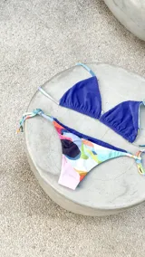 Bikini reversible de diseño triangular con tiras para atar y apliques de argollas metálicas. El conjunto presenta un estampado abstracto multicolor.
