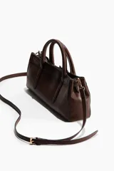 Cartera bandolera rectangular de color marrón, con diseño alargado y asas cortas de mano. Incluye un detalle de tira decorativa con hebillas metálicas en la parte frontal y herrajes dorados.