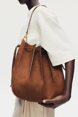 Cartera bandolera de gamuza color marrón, con diseño de pliegues cosidos y cordón ajustable en la parte superior. Cuenta con correa de hombro ajustable y herrajes metálicos.