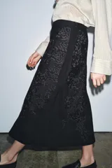 Falda larga negra de tiro medio confeccionada en hilatura con mezcla de algodón. Detalle de bordados florales a tono. Bajo evasé y cierre con cremallera oculta en costura.