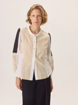 Camisa de manga larga con diseño patchwork que combina paneles de tela texturizada en tonos beige, blanco y negro. Presenta cuello redondo, cierre frontal con botones y un corte holgado.