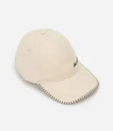 Gorra de algodón color beige con visera curva. Presenta bordado frontal en cursiva negra con la frase "less is more" y pespuntes contrastantes en negro a lo largo del borde de la visera. Cuenta con cierre ajustable en la parte posterior mediante hebilla metálica.