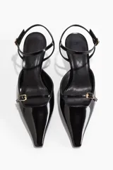 Sandalia negra de charol con punta fina, taco kitten y pulsera ajustable al tobillo. Presenta una tira delantera con hebilla dorada.