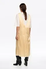 Vestido midi de diseño bimateria con sobrevestido de satén color amarillo claro. Presenta escote en V profundo con lazo frontal, detalles de encaje en el ruedo y aberturas laterales con cordones. Se combina con una base de remera blanca de manga corta y cuello redondo.