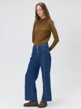 Jeans de corte ancho (wide leg) y tiro medio, confeccionados en denim de algodón negro con costuras contrastantes en color ocre. Presenta múltiples botones metálicos frontales y tachas metálicas aplicadas a lo largo de los laterales y la pretina.