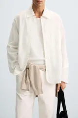 Camisa de corte relajado, color blanco hueso, con finas rayas verticales en tono grisáceo. Presenta cuello solapa, manga larga, bolsillo de parche en el pecho y bajo con aberturas laterales.