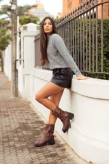 Bota corta de cuero color marrón, con taco cuadrado y punta fina. Presenta una correa con tachas pequeñas y una argolla metálica decorativa alrededor del tobillo. Cuenta con cierre lateral.