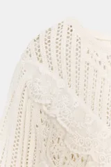 Jersey de punto color blanco roto, manga larga, con cuello redondo y detalle de encaje en el delantero en forma de volados en 'V'. El bajo es asimétrico.