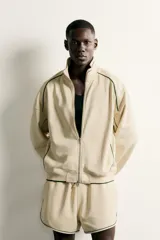 Chaqueta estilo deportivo de corte relajado, color beige con detalles de vivos en color verde oscuro. Presenta cuello alto, cierre frontal con cremallera de doble dirección, bolsillos laterales y puños elásticos.