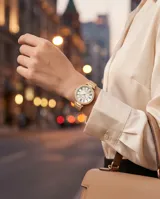 Reloj de pulsera analógico con caja redonda de metal dorado y bisel con incrustaciones de cristales. Esfera blanca con números romanos y malla de acero tipo milanesa en tono dorado.