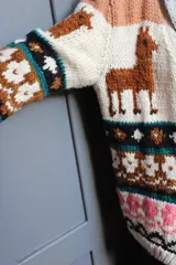 Cardigan tejido a mano en lana merino, con diseño multicolor de inspiración andina que incluye motivos de llamas, franjas geométricas y detalles en tonos beige, marrón, negro y rosa.