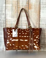 Cartera tipo tote de cuero con frente de piel con estampado animal print tipo ciervo en tonos marrón y blanco. Posee manijas de cuero marrón con hebillas metálicas decorativas y flecos colgantes en uno de los laterales.