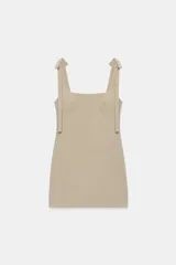 Vestido corto de punto acanalado en color beige, con escote cuadrado y tirantes con detalle de lazos. Presenta un diseño ajustado y cierre posterior con cremallera oculta.
