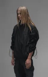 Campera rompevientos negra de calce oversize, confeccionada en microfibra técnica ligera con acabado semi-mate. Presenta capucha integrada, cierre frontal técnico y bolsillos tipo ojal. Destaca por su sistema de frunces elásticos regulables mediante tancas a lo largo de las mangas y el ruedo, permitiendo ajustar el volumen de la prenda.