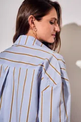 Camisa de corte oversize con estampado de rayas verticales en tonos azul claro y mostaza. Presenta cuello clásico, cierre frontal con botones y mangas largas con puños abotonados.