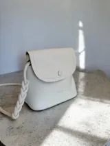 Cartera tipo bandolera de color blanco con textura granulada, solapa con cierre de botón magnético y correa trenzada ajustable mediante nudos.