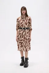 Vestido tipo camisa de corte holgado, con cuello clásico, cierre frontal de botones y mangas largas. Presenta un estampado de hojas en tonos marrones y beige.