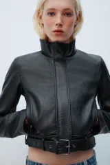 Cazadora de cuero sintético negro con textura granulada, cuello alto, manga larga y cierre frontal con cremallera oculta. Cuenta con bolsillos delanteros y un cinturón ajustable con hebilla metálica en el bajo.