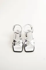 Sandalias de cuero color plateado con taco ancho y bajo. Presentan tiras cruzadas sobre el empeine y una tira fina con hebilla ajustable en el tobillo. La punta es cuadrada.