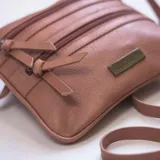 Bandolera color marrón con apertura superior de cierre metálico y dos bolsillos frontales con cierre. Incluye correa larga regulable y placa metálica con el logo de la marca.