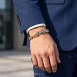 Pulsera de eslabones gruesos con baño de oro, destacada por un cierre central de diseño ornamental en forma de flor de lis con acabado plateado.